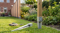 Galvanised drive-in fence post spikes on a garden lawn: one driven in holding a square timber post, a second lying ready beside it