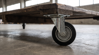 Solid rubber swivel castor bolted under a heavy timber workshop trolley on a concrete warehouse floor