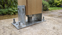 Hot-dip galvanised height-adjustable post support bolted to a concrete slab, with the central threaded rod and adjusting nut visible beneath a square timber post held clear of the surface