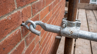 Galvanised scaffold wall tie: a hooked anchor tube engaging an eye bolt fixed into red brickwork, clamped to a vertical scaffold standard with a right angle coupler.